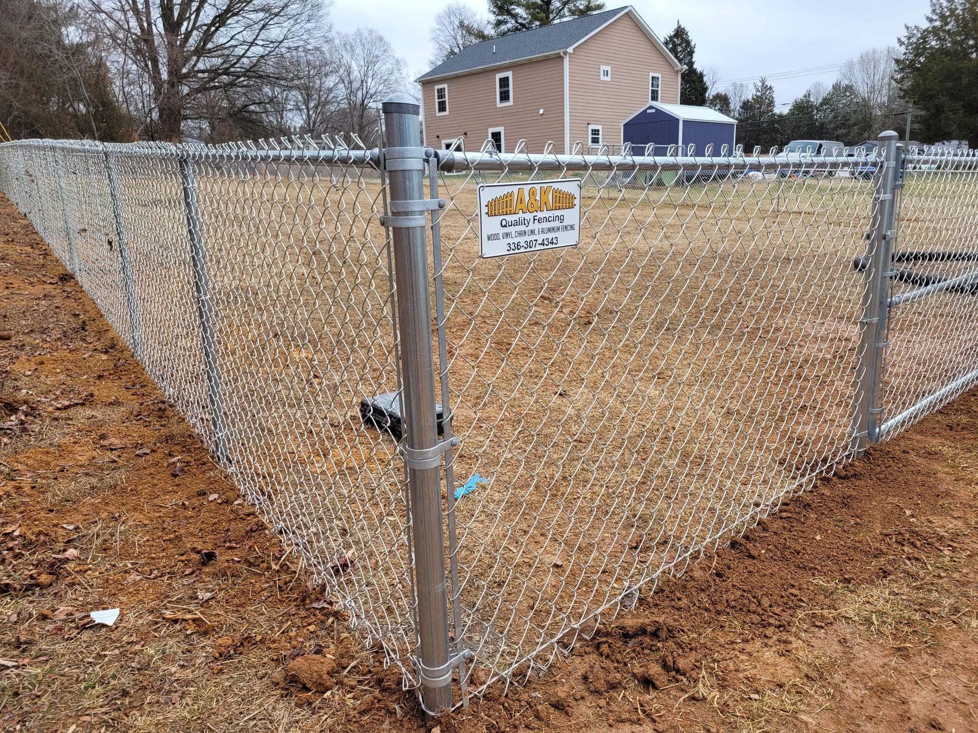 Chain-link fence in front of a vacant lot; a house is in the background. A sign hangs on the fence.