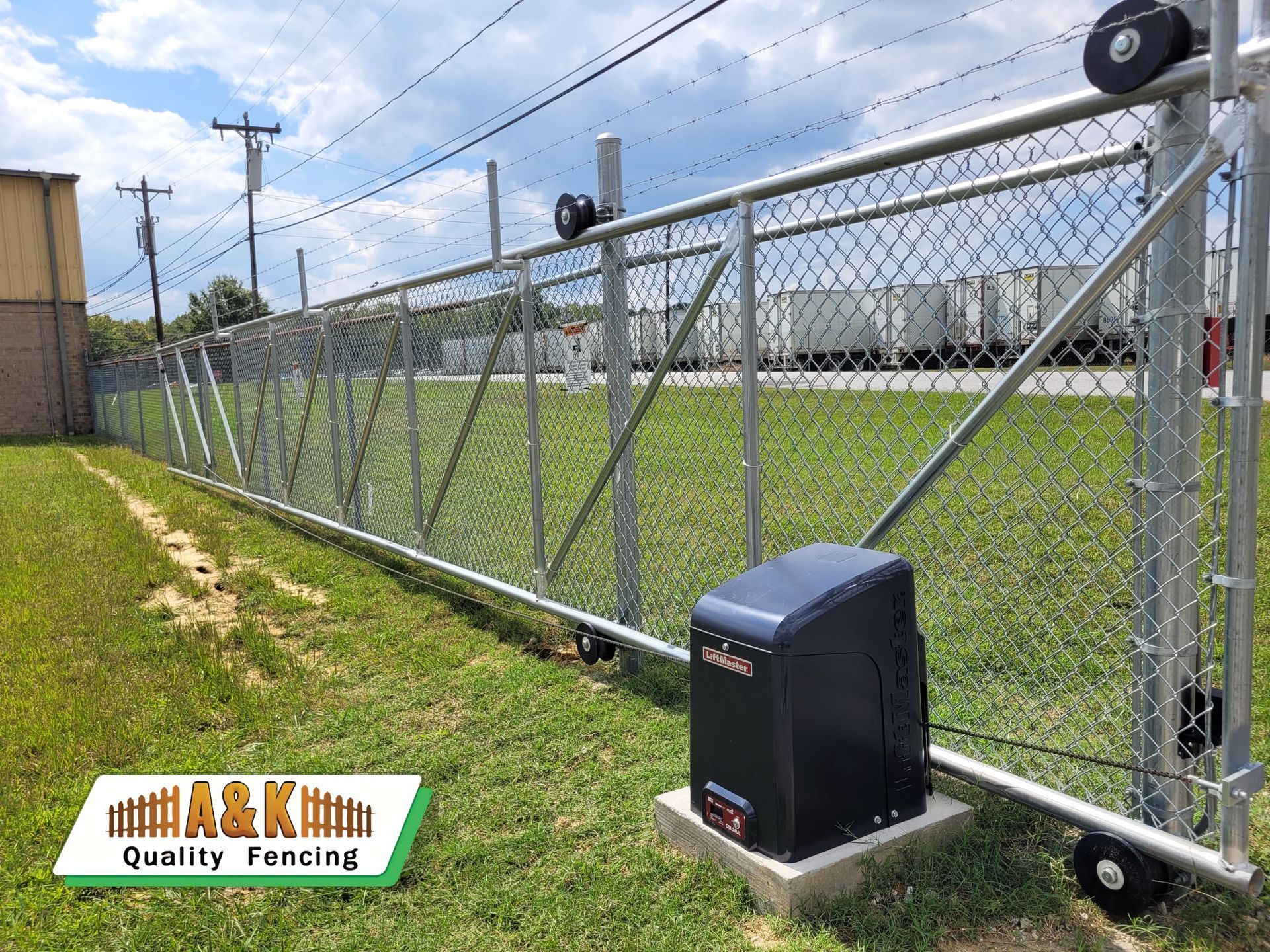 Chain-link fence with an automatic gate opener installed, in front of a building and road.