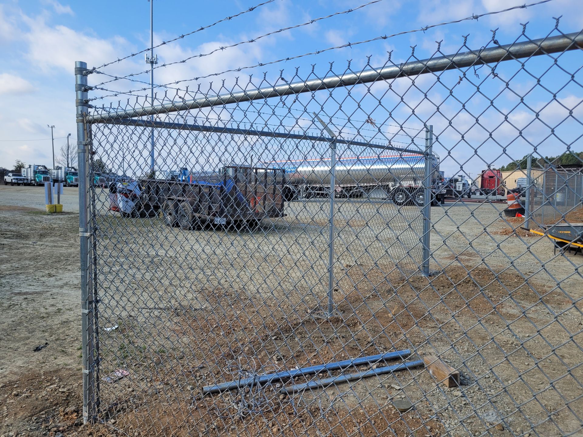 Chain-link fence topped with barbed wire encloses an industrial lot with vehicles and buildings under a cloudy sky.