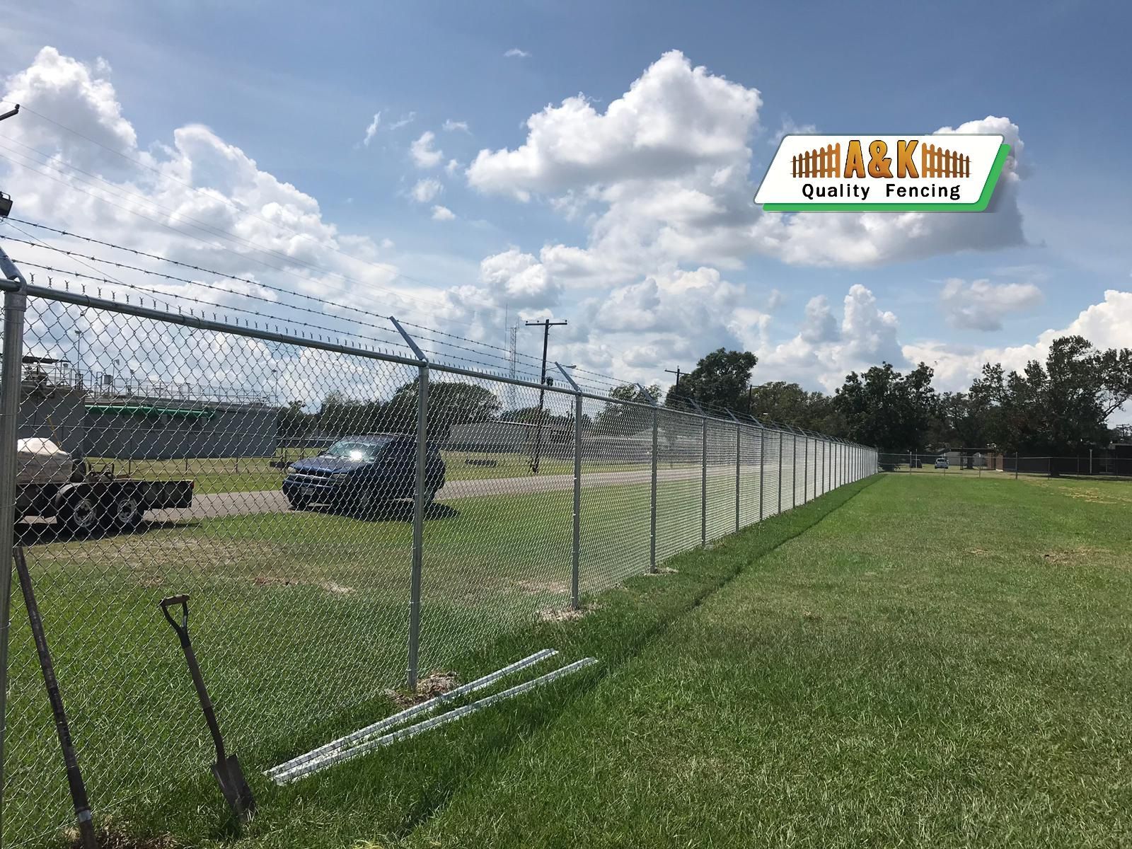 Chain-link fence with barbed wire on top, enclosing a grassy area, a vehicle, and equipment, under a blue sky.