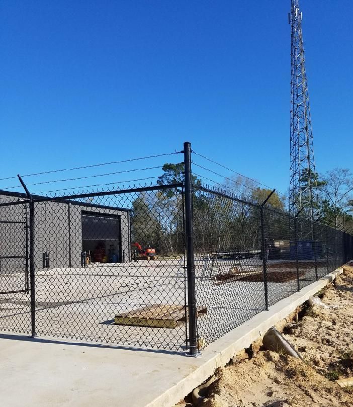 Black chain-link fence with barbed wire surrounds a building and radio tower under a clear blue sky.