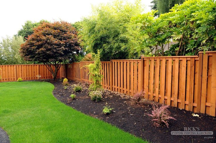 Wooden fence lines a lush green lawn and garden bed with various plants and trees.