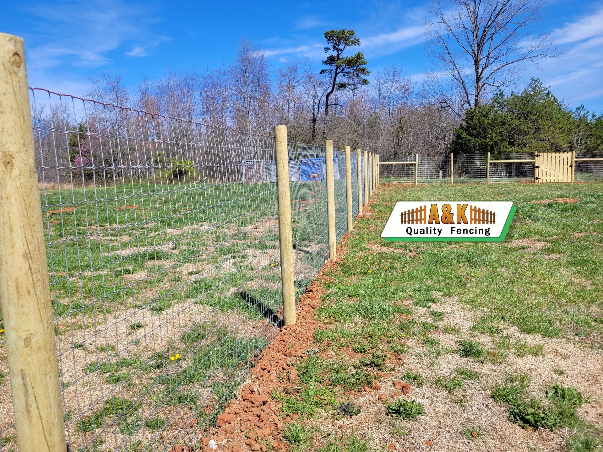 A wire fence with wooden posts in a field, under a blue sky, with some trees and a small sign.