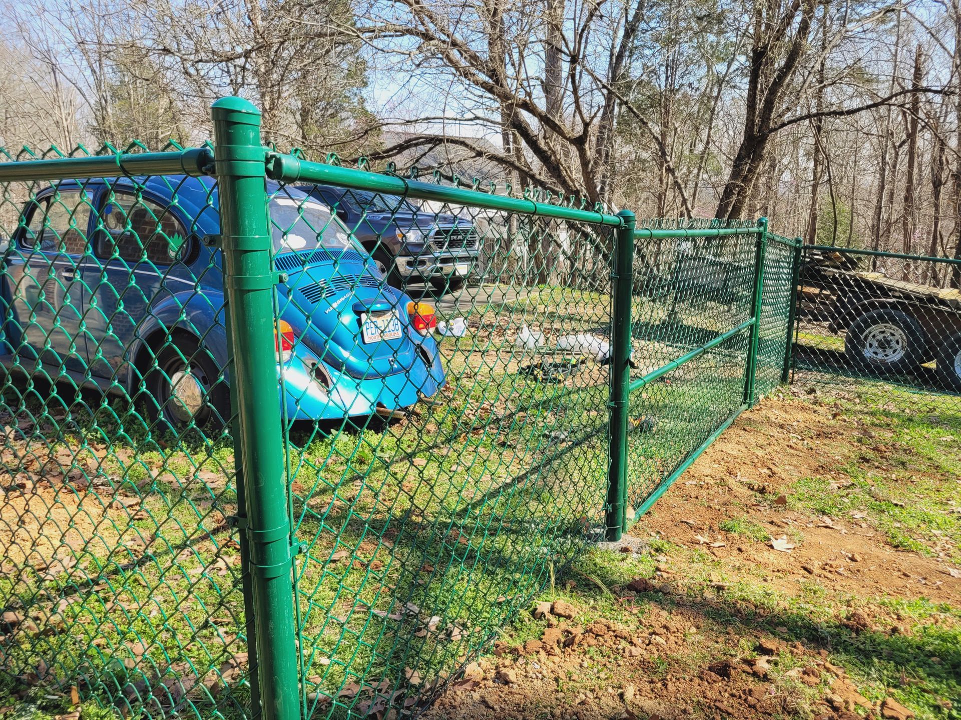 Green chain-link fence surrounds a grassy area with a blue car and a truck parked behind it. Trees in the background.