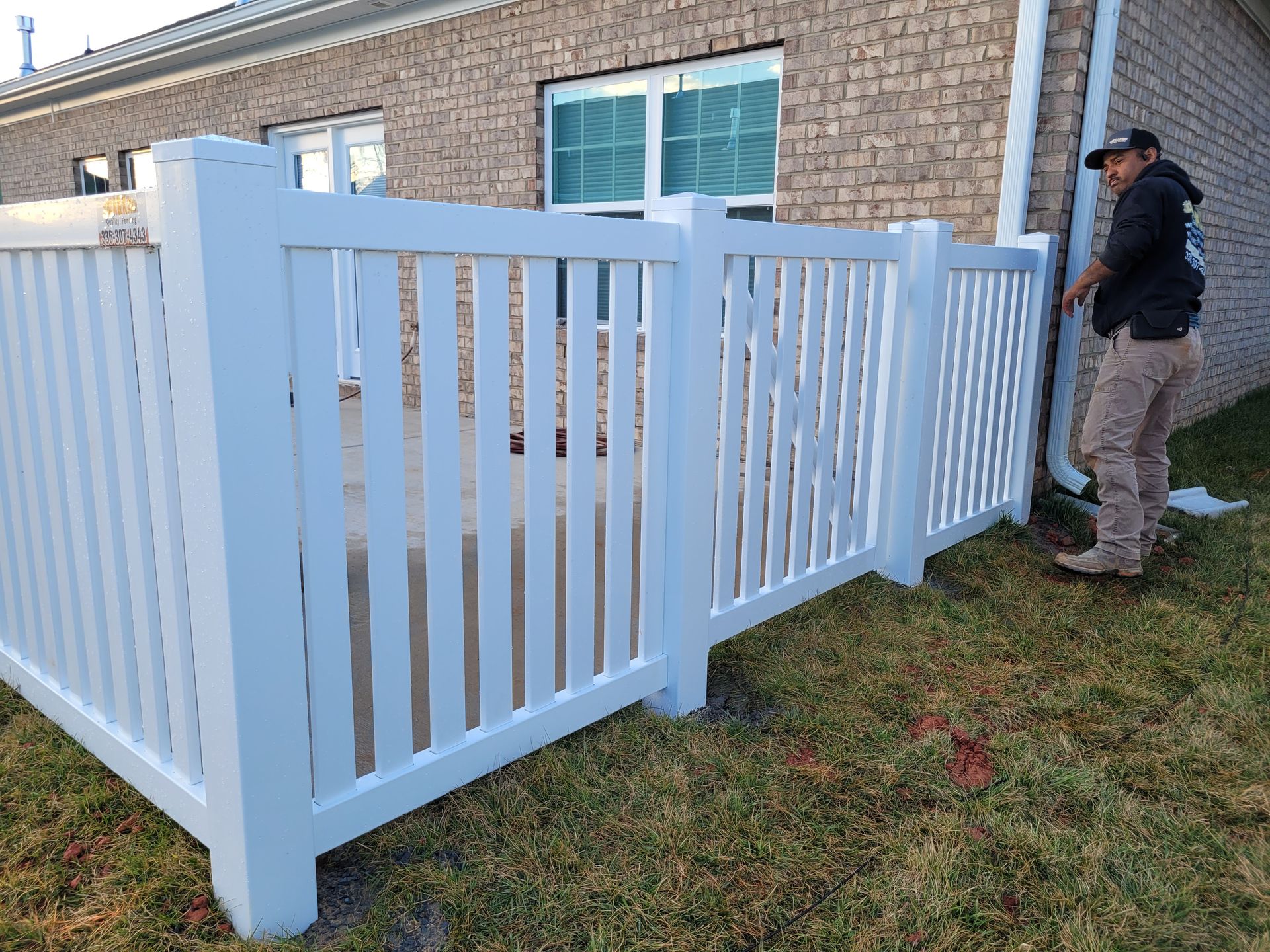 Man installing a white picket fence next to a brick building and a grassy area.