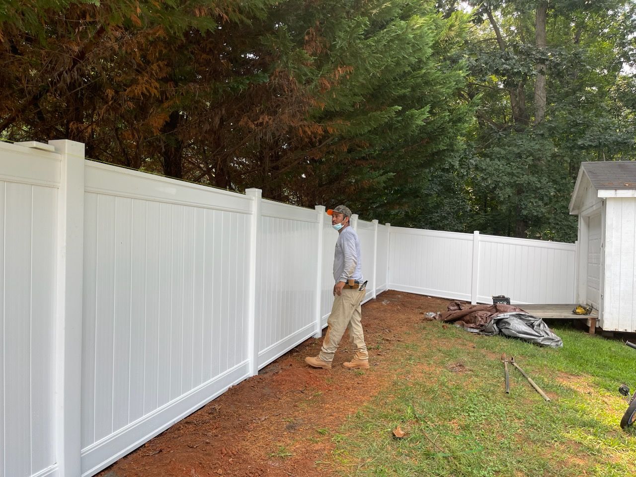 Man walking alongside newly installed white vinyl fence; yard with shed and trees.