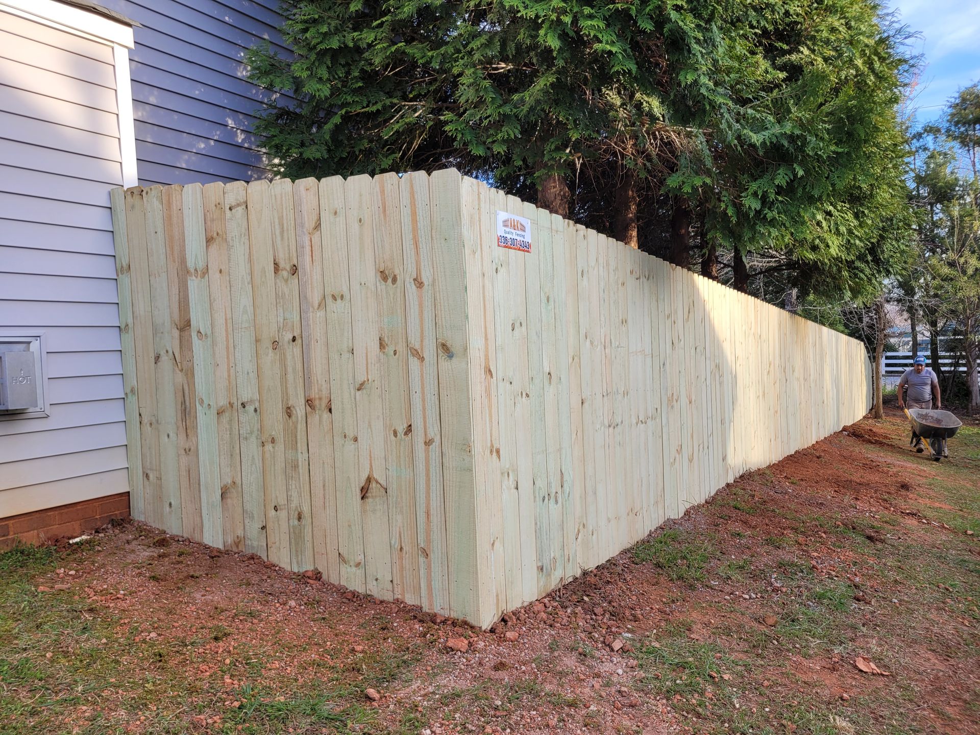 Wooden fence bordering a yard next to a gray house, with a tree in the background.