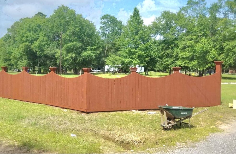Red wooden fence with decorative tops, green grass, trees, and a wheelbarrow.