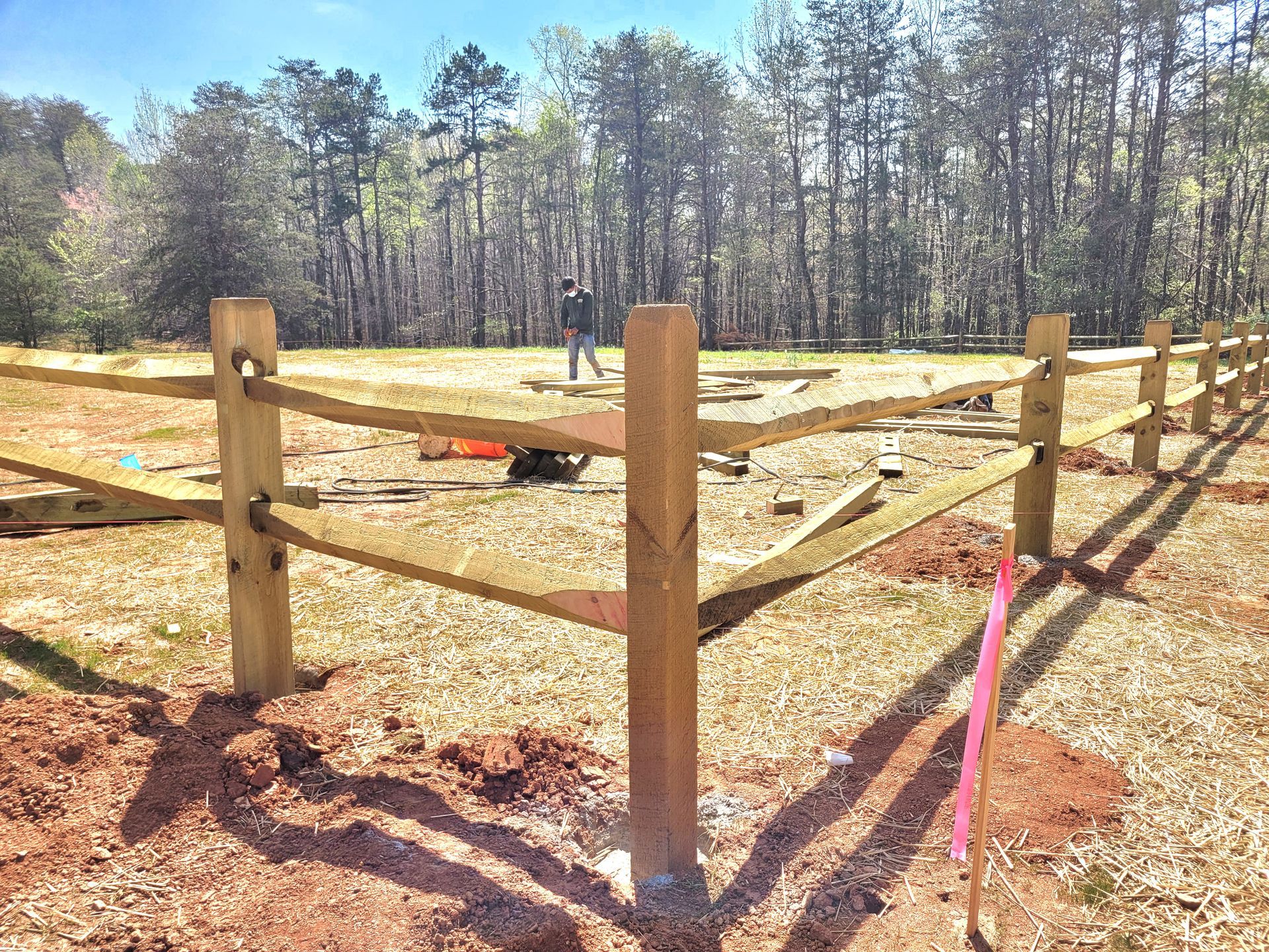 Wooden fence being constructed in a field with a treeline in the background. Construction worker in the distance.