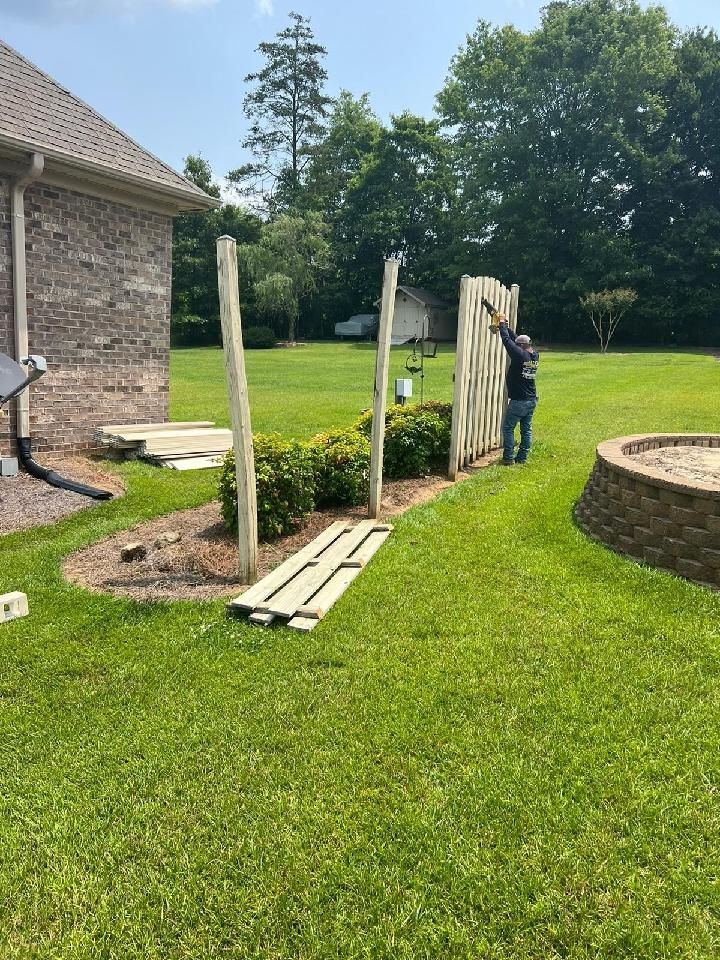 Man installing a fence in a backyard, with a brick house and fire pit visible.