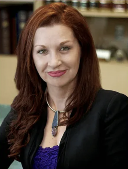Portrait of a woman with red hair in a black blazer and purple top, seated indoors and smiling at the camera
