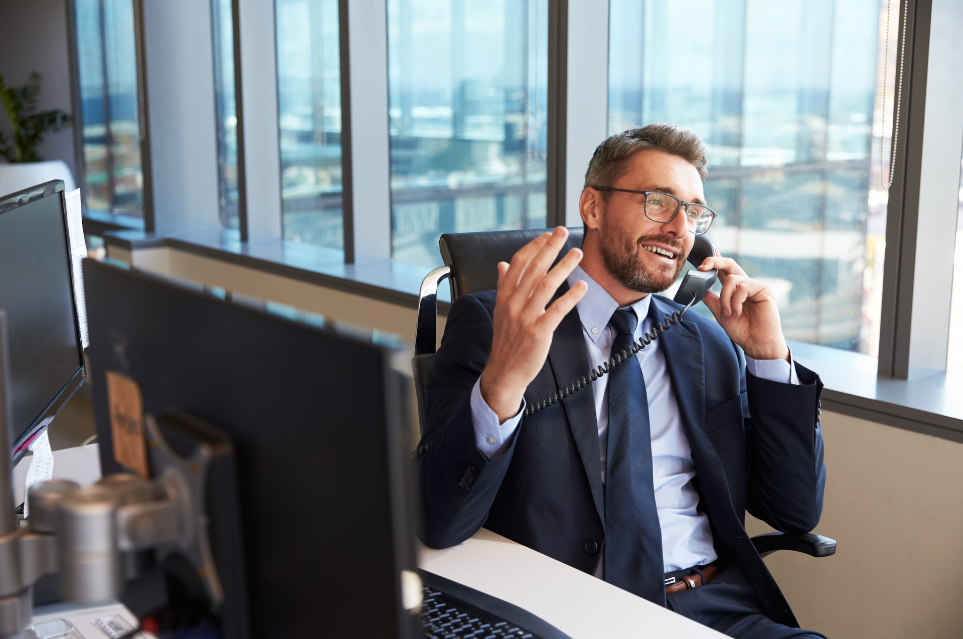 A man in a suit and tie is sitting at a desk talking on a phone.