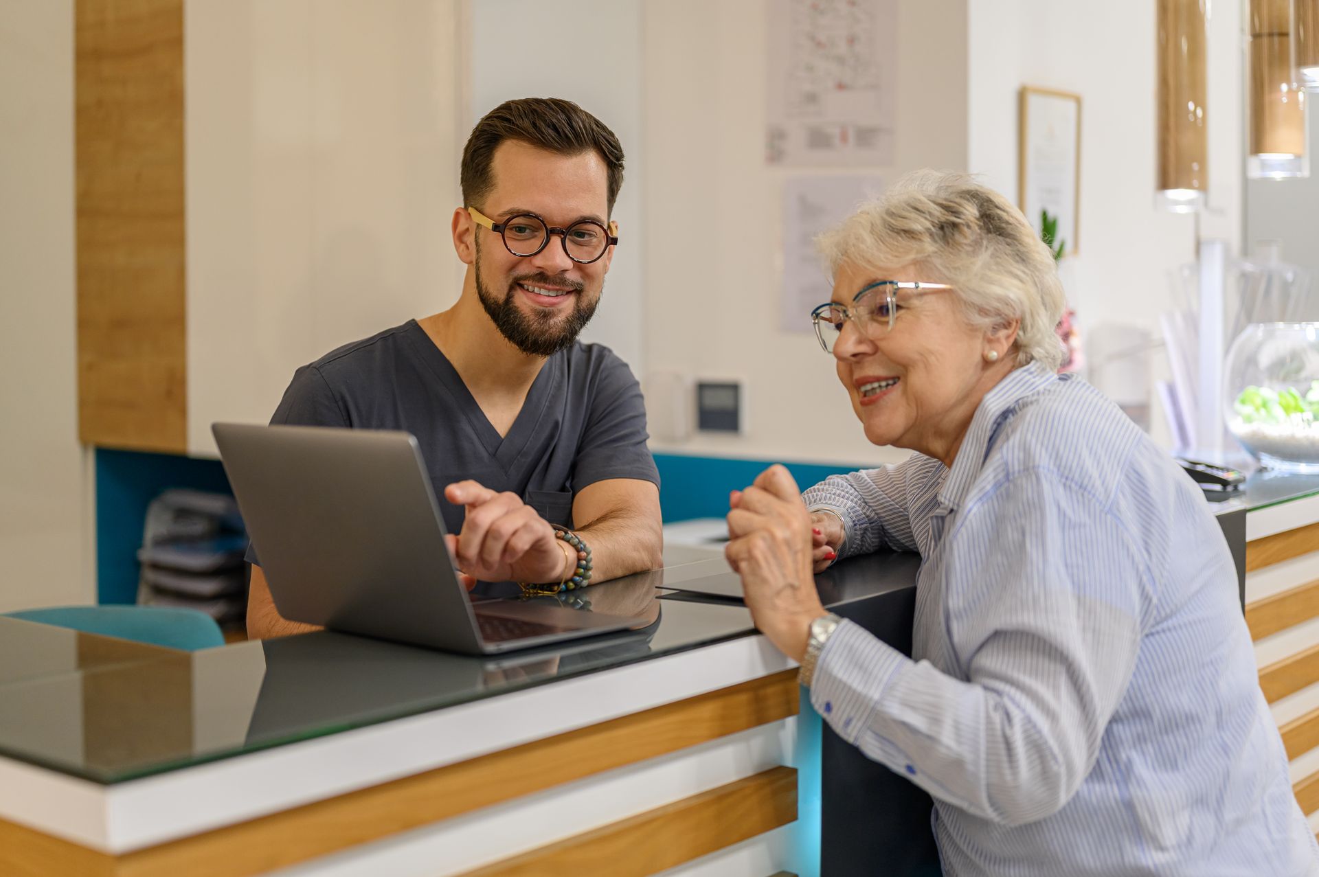 An eye doctor and senior woman collaborate at a clinic desk, focused on a laptop in front of them. An eye doctor and senior woman collaborate at a clinic desk, focused on a laptop in front of them.