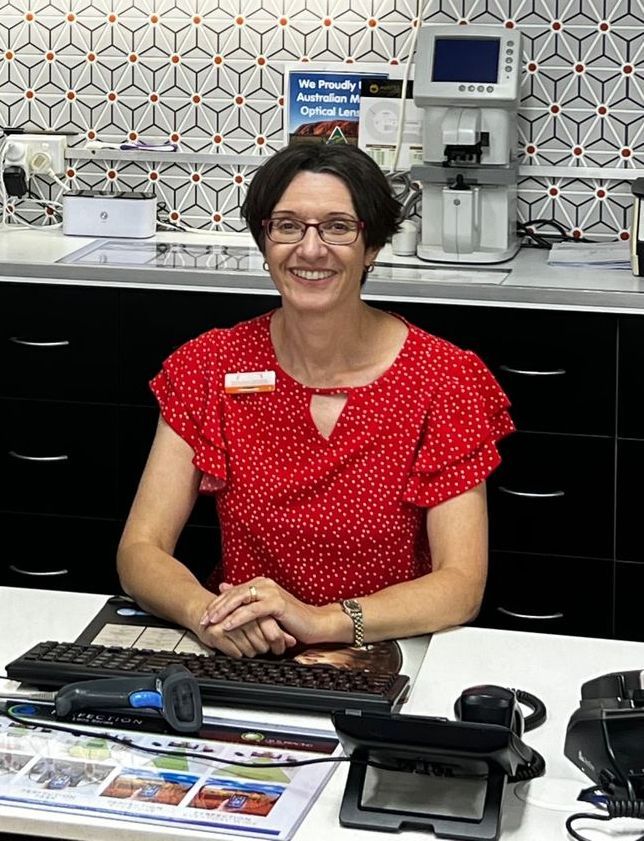 Woman in red dotted shirt smiles, seated at a desk, office setting with eye exam equipment in background.