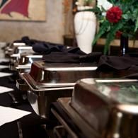 Polished silver serving trays lined up on a buffet table with black cloth napkins and red flowers in the background