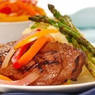 Grilled steak topped with colorful peppers and onions, served with asparagus on a white plate