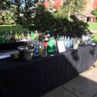 Outdoor drink station with bottles, cups, and ice buckets on a black-covered table in a sunny courtyard