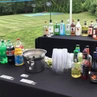 Outdoor drink station with assorted bottles, stacked plastic cups, and an ice bucket on black tables