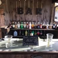Bar setup with bottles, a chalkboard drink sign, and plastic cups on a rustic wooden counter