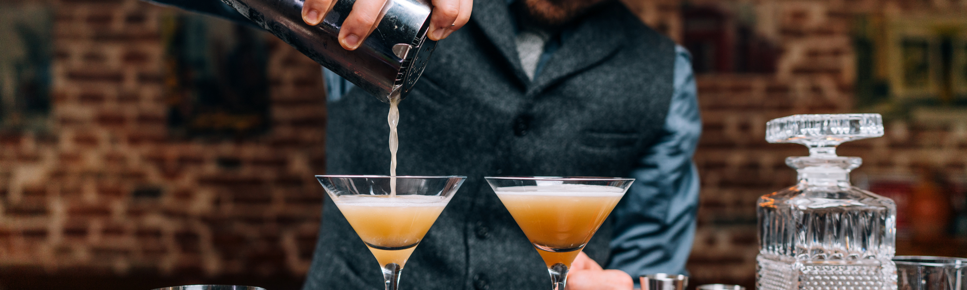 Bartender pouring liquor into a martini glass beside two cocktails on a bar counter