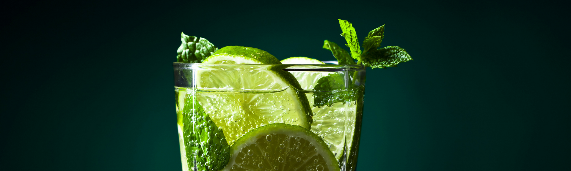 Glass of lime drink with ice, mint, and lime slices against a dark green background