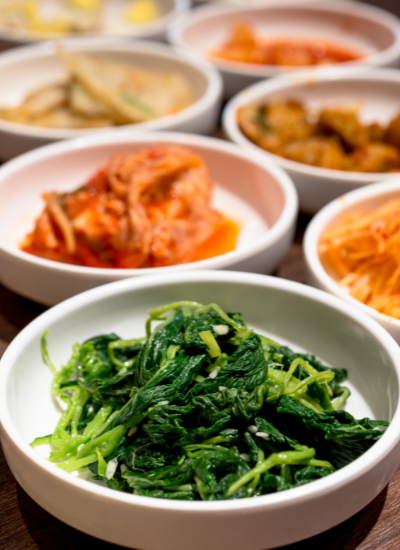 Bowls of assorted Korean side dishes, with a bowl of green seasoned spinach in front