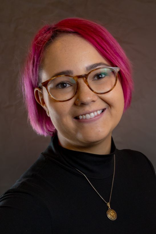 A woman with chin length pink hair and larger round glasses smiling.