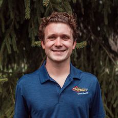 A young man in a blue shirt is smiling in front of a tree.