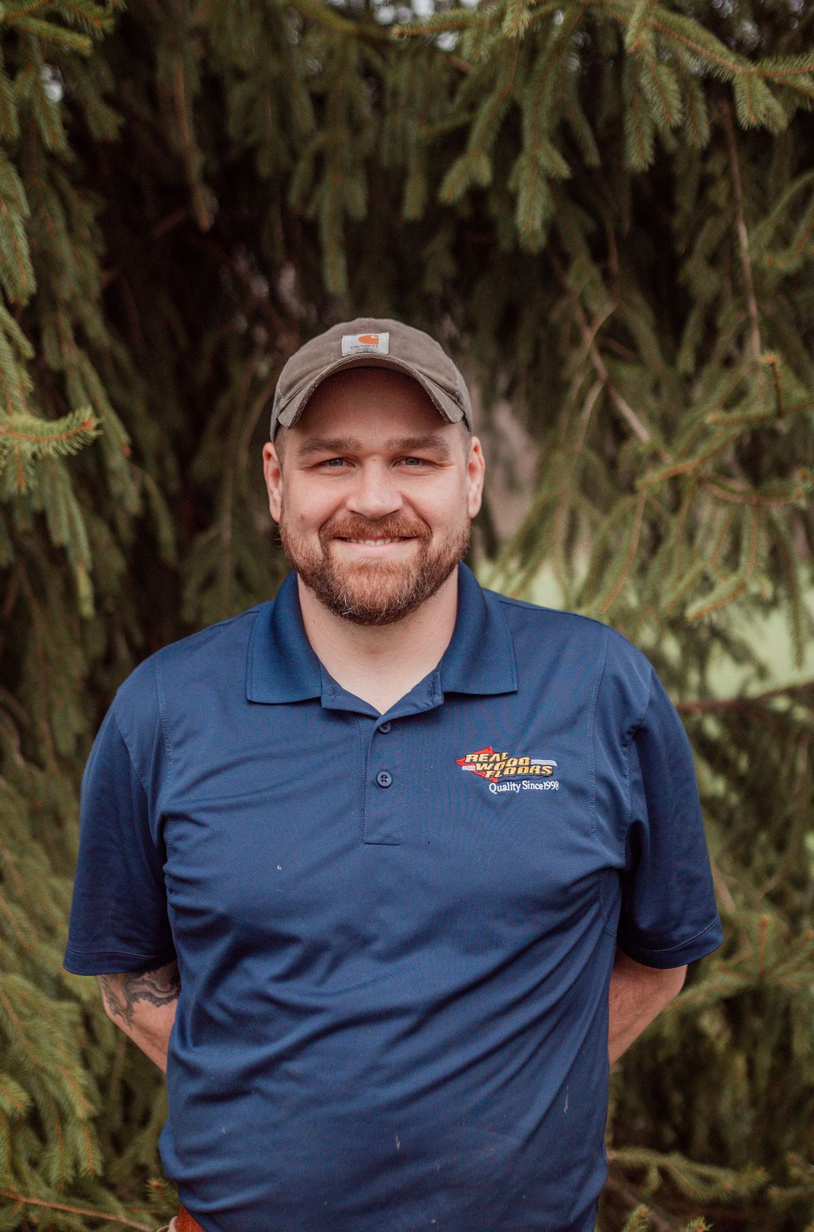 A man in a blue shirt and hat is standing in front of a tree.