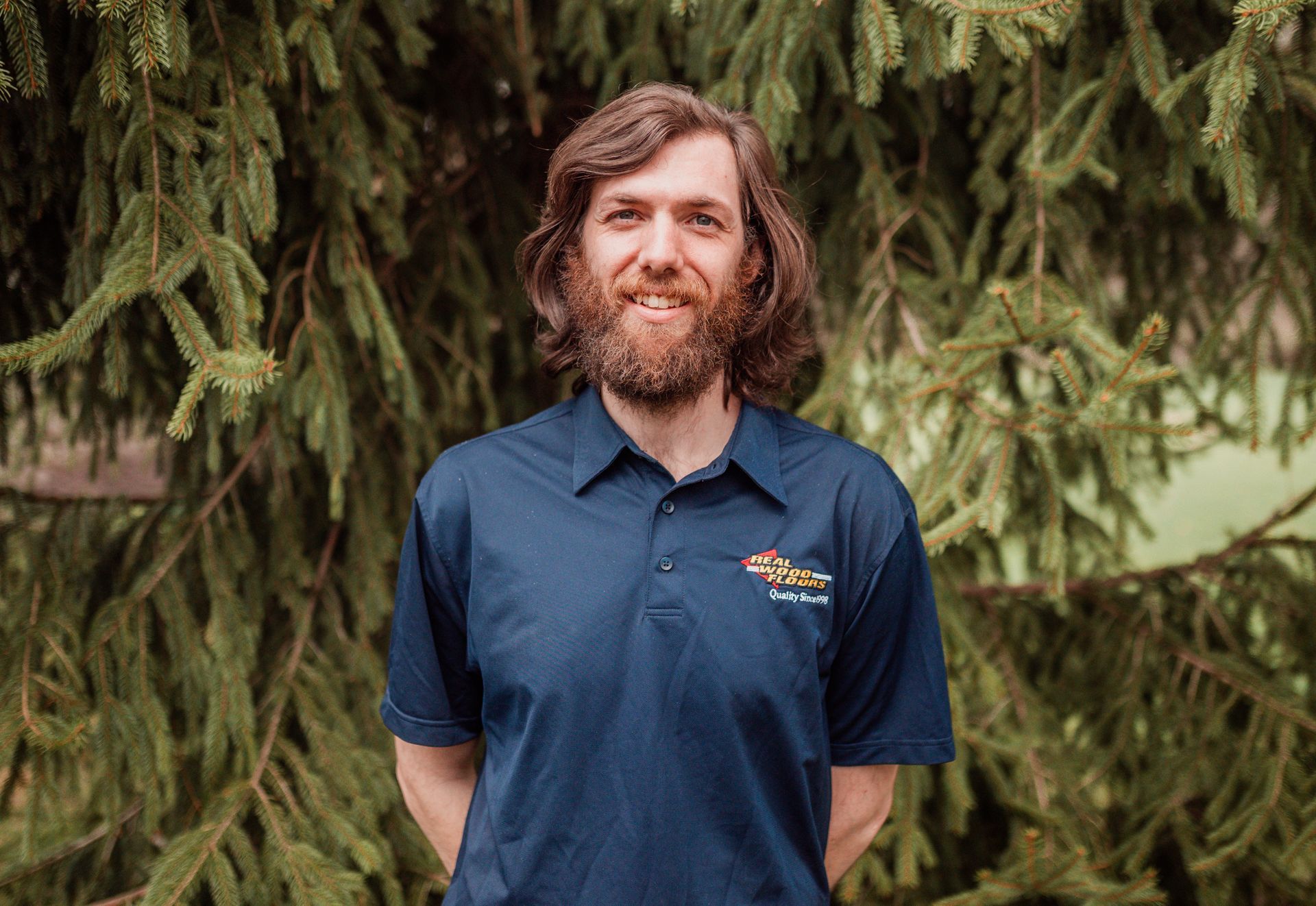 A man with a beard wearing a blue polo shirt is standing in front of a tree.