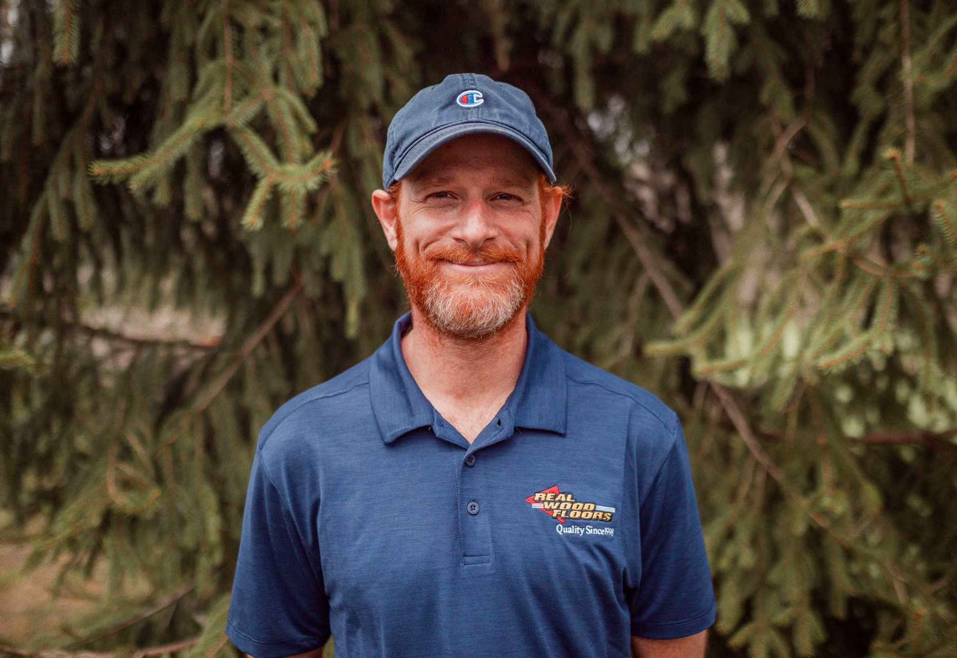 A man with a beard and a hat is standing in front of a tree.