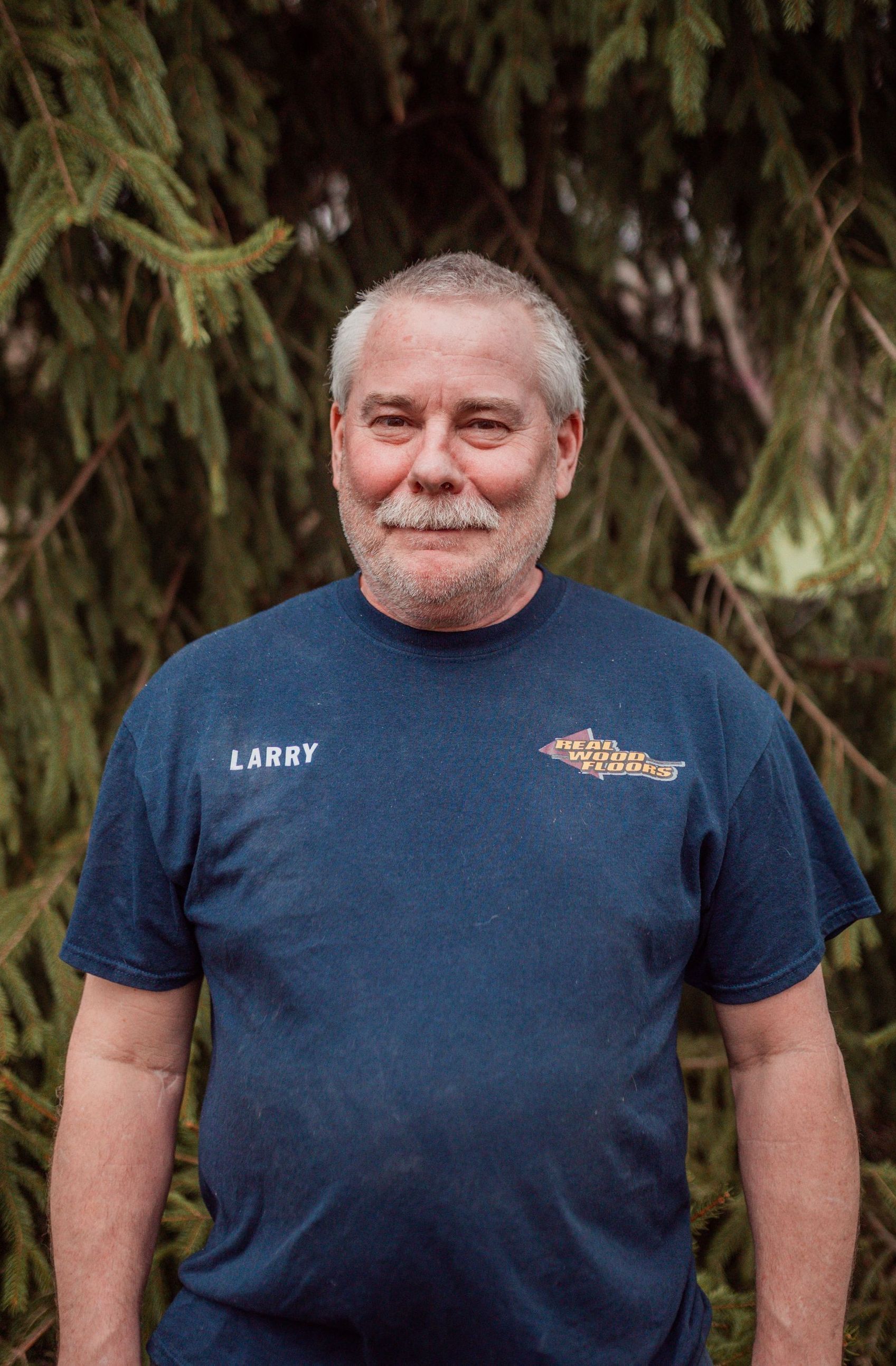 A man with a beard wearing a blue t-shirt is standing in front of a tree.