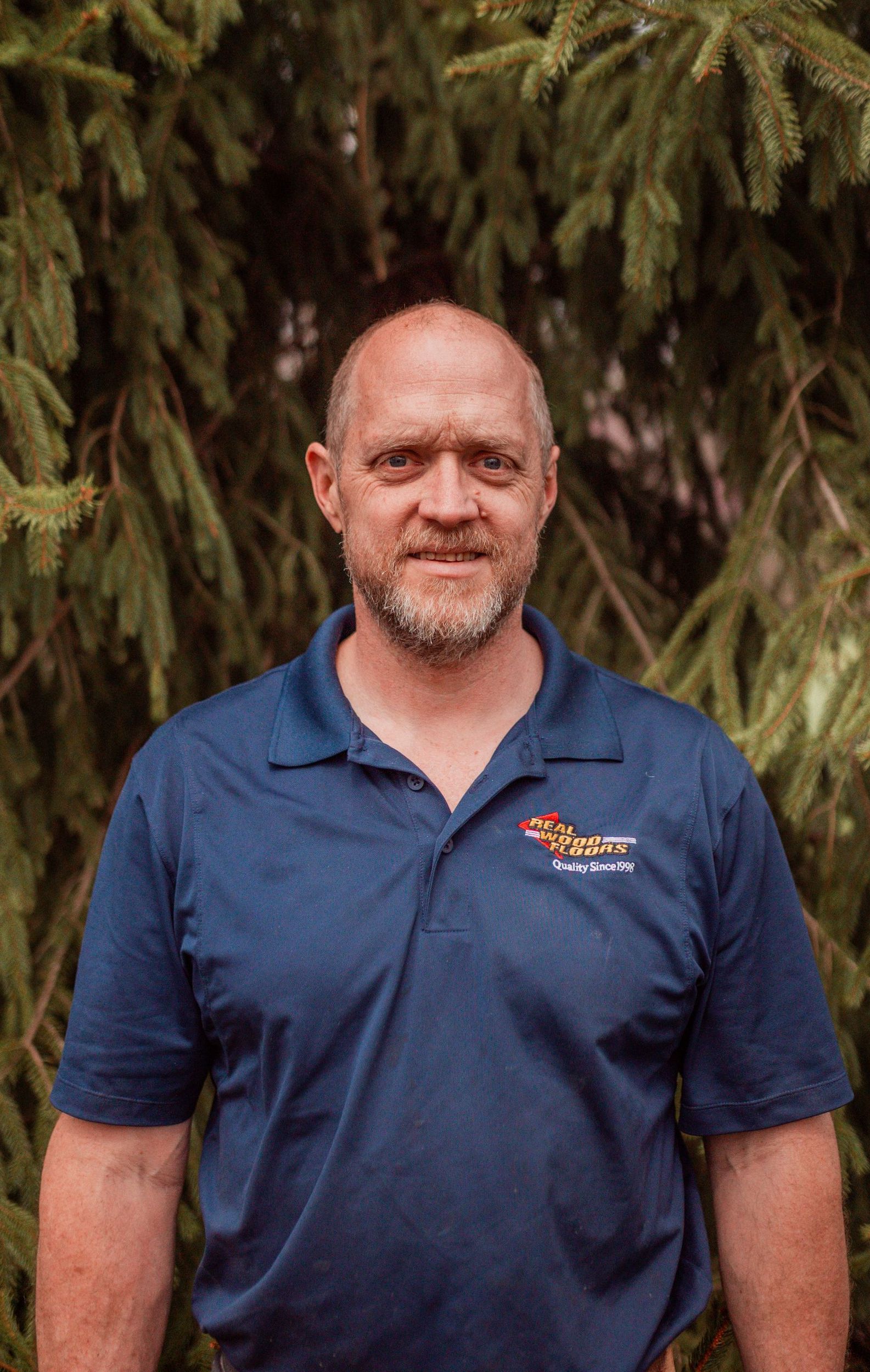 A man with a beard is wearing a blue shirt and standing in front of a tree.