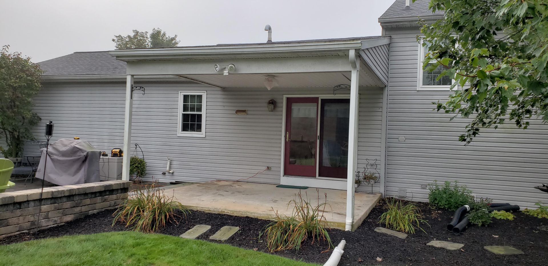 A gray-sided house exterior featuring a covered patio, a sliding glass door, and a landscape garden with mulch.