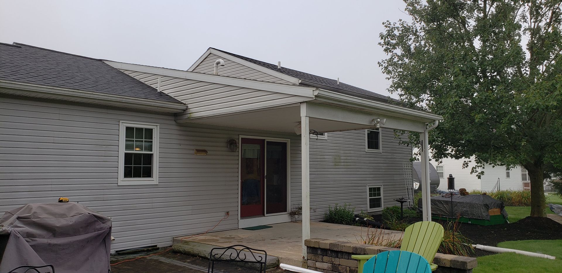 Exterior view of a house with light gray siding, a covered patio area, and lawn chairs on a cloudy day.