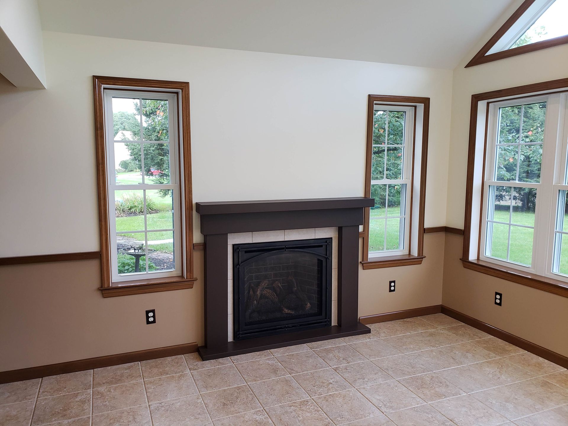 A bright room features a dark fireplace mantel centered between two windows, with tan wainscoting and tiled flooring.