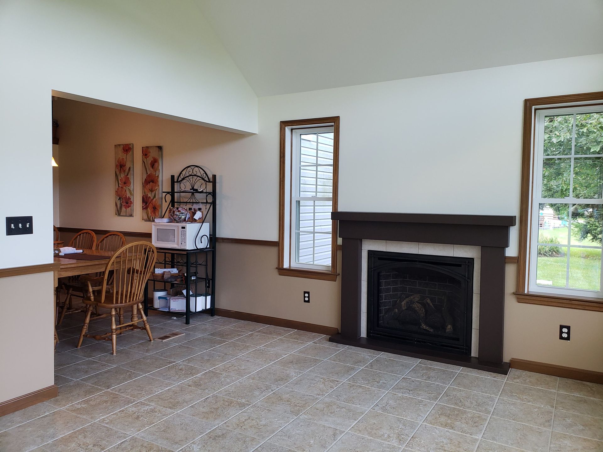 A living room with a fireplace, beige and white walls, tiled floor, and a view into a dining area with a metal shelving unit.