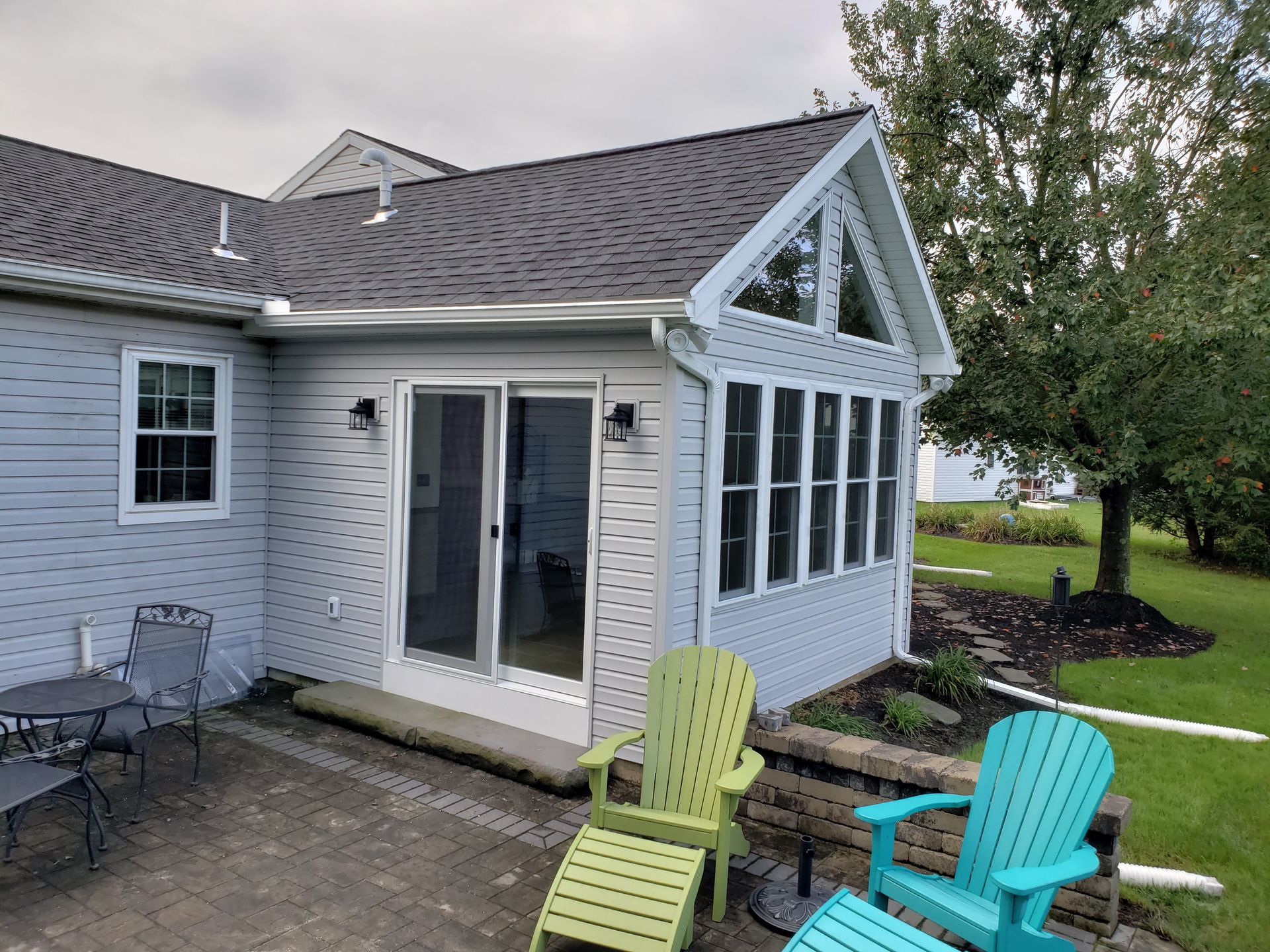 A sunroom addition with light blue siding and a slate roof, featuring a patio with colorful chairs and a stone wall.