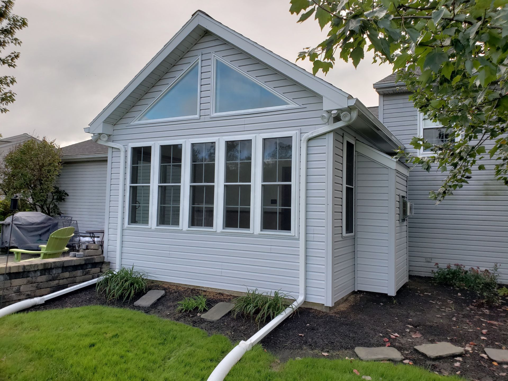 A light gray house with a triangular window above four rectangular windows and white downspouts in a grassy yard.