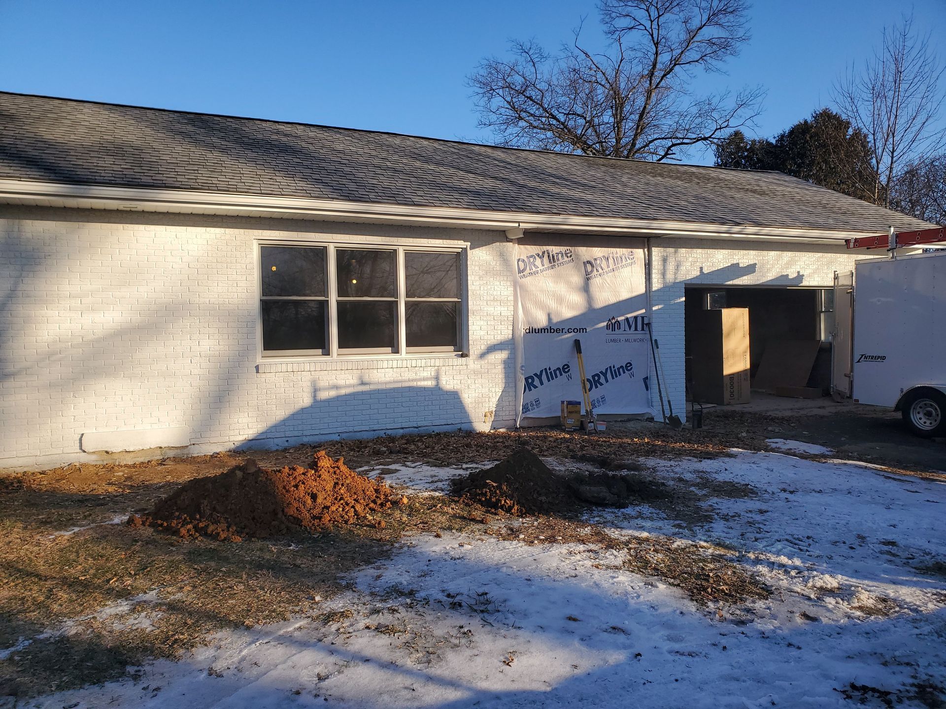 A white brick house under renovation with a pile of dirt in the foreground and a utility trailer parked on the side.