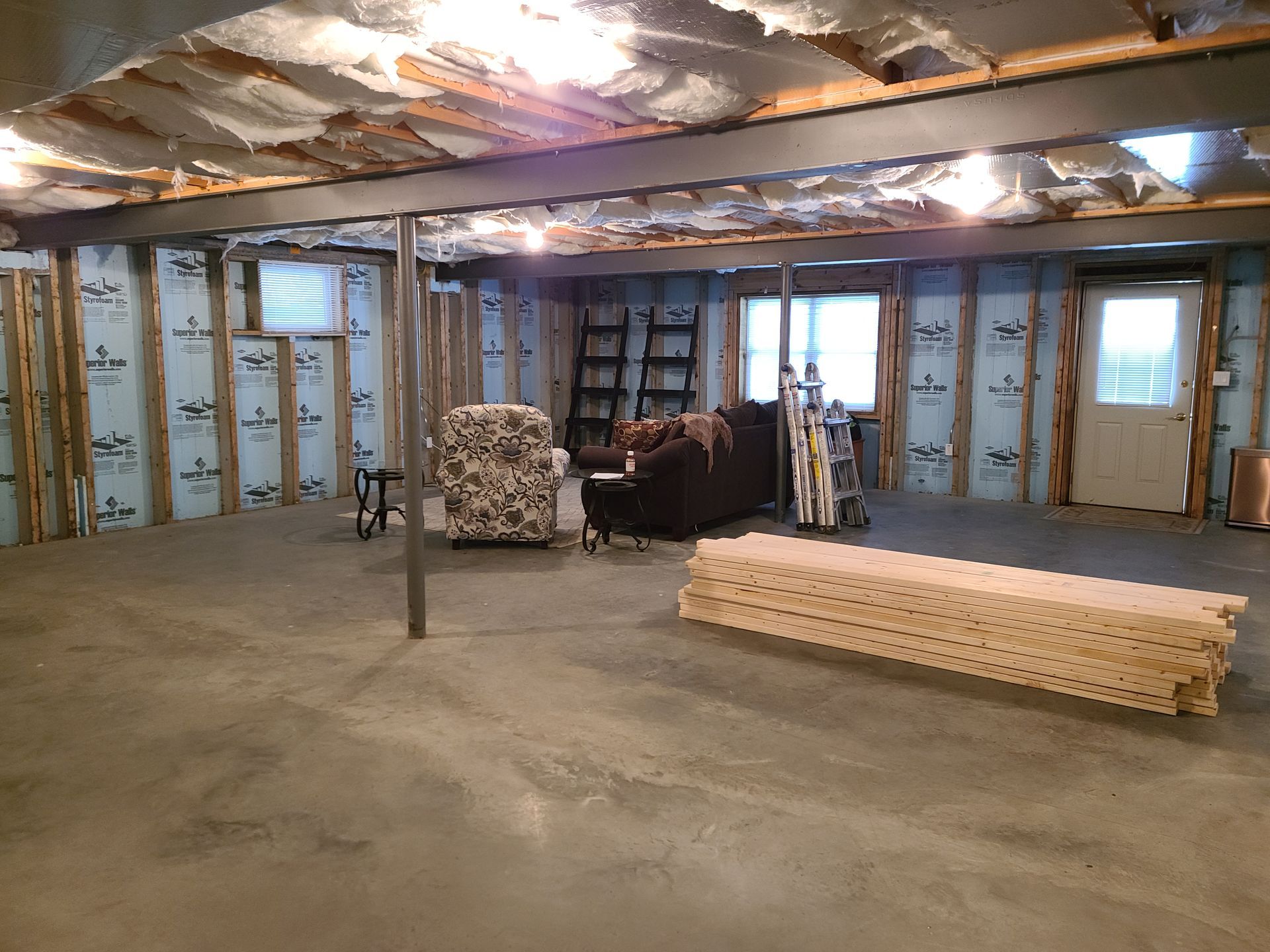 A partially finished basement with concrete floors, exposed ceiling beams, insulated walls, and a small seating area.