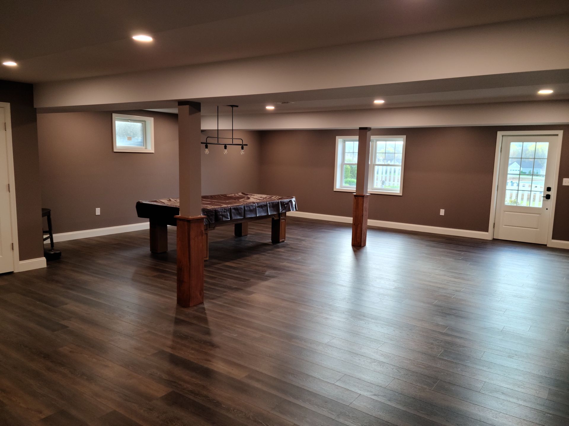 A spacious basement with dark brown walls, wood-look flooring, two support pillars, a pool table, and an exterior door.