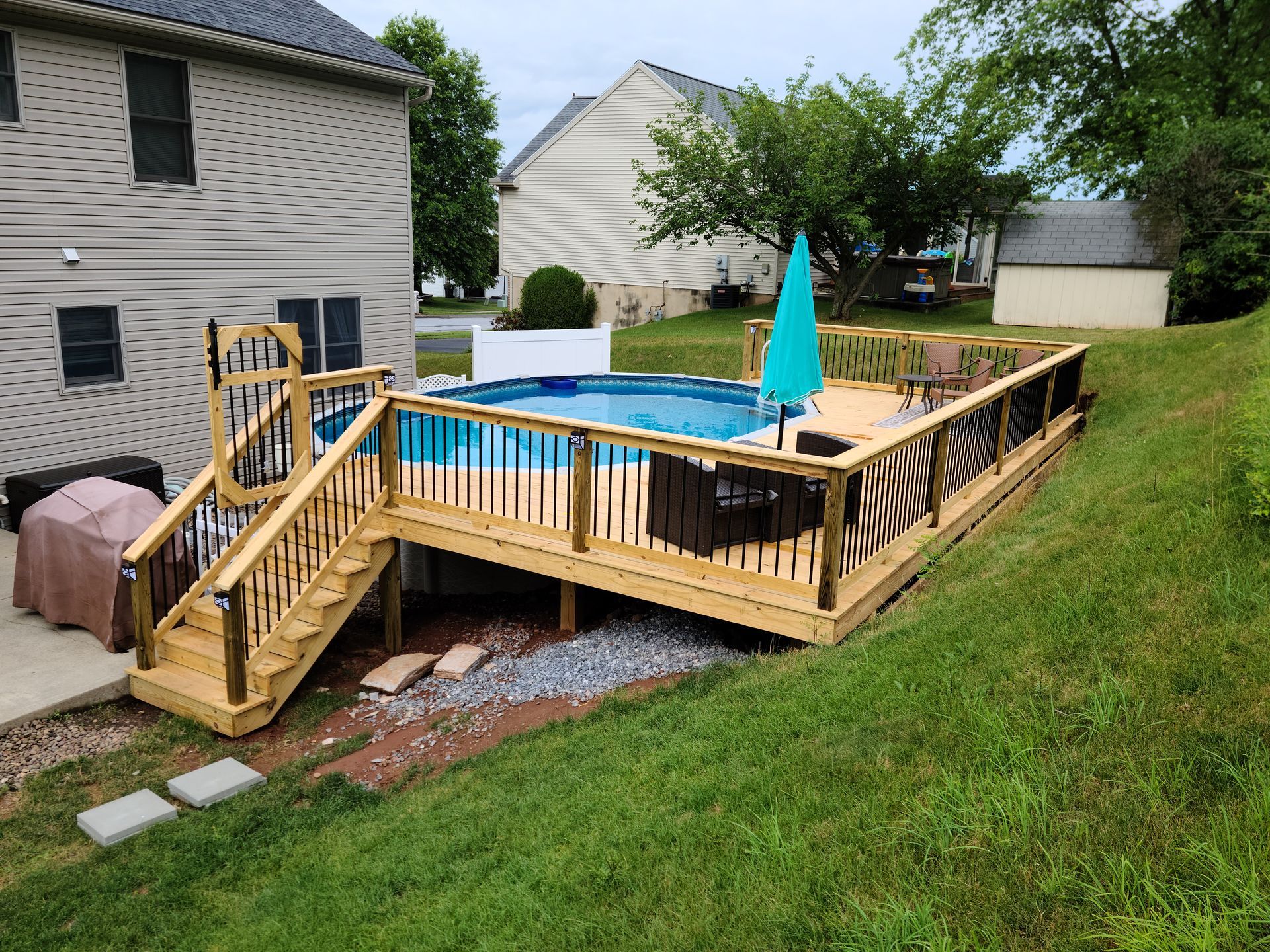 An above-ground swimming pool with a wraparound wooden deck and stairs, situated in a backyard beside a house.