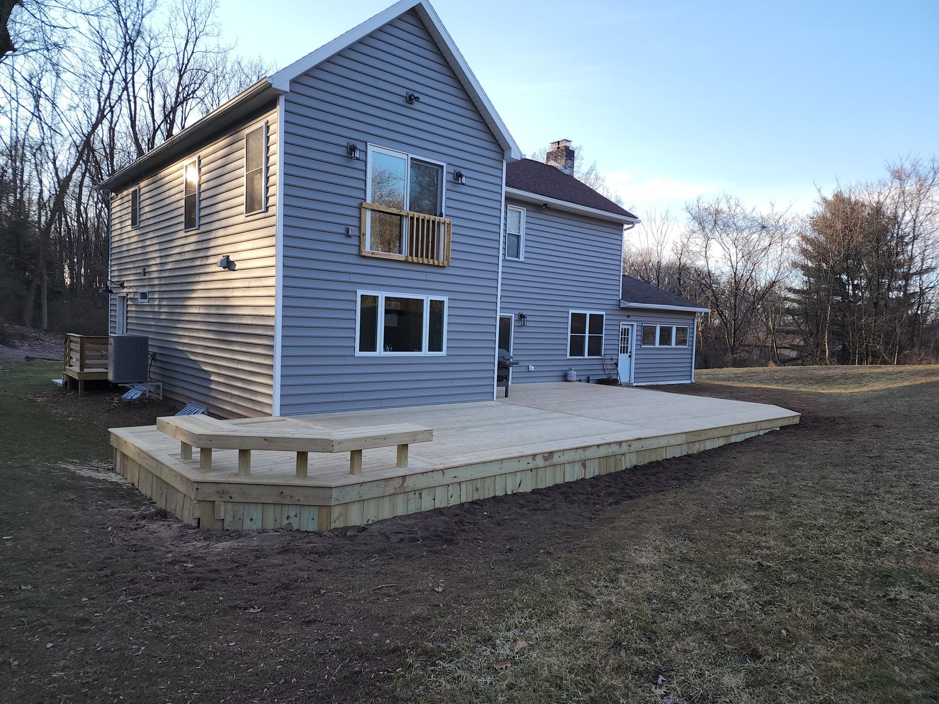 A gray, two-story house with a newly constructed wooden deck featuring a built-in bench in a grassy, rural backyard.