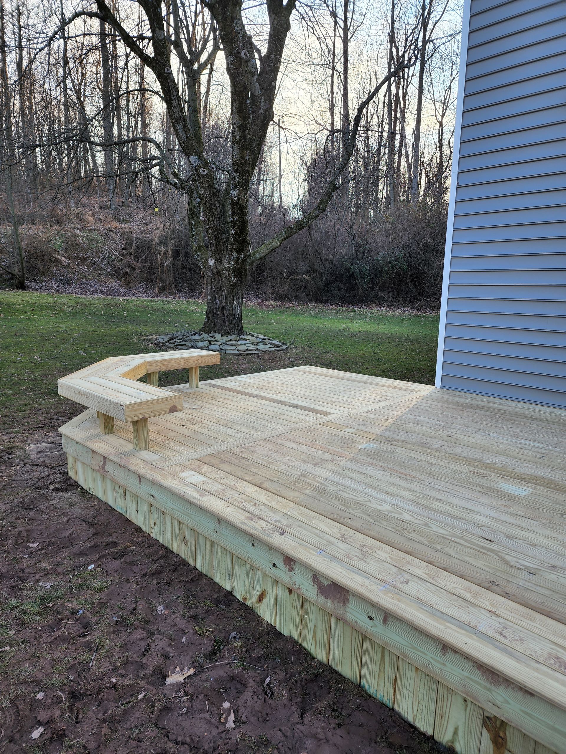 A newly built wooden deck featuring a curved, built-in bench, situated on a grassy lawn next to a grey-sided house.