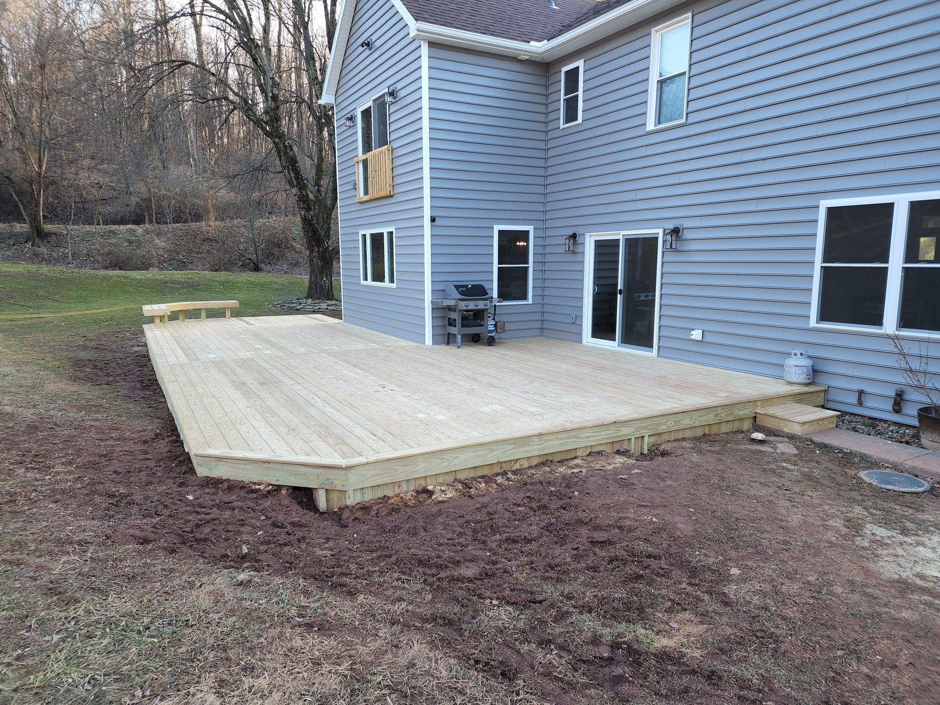 A new light-wood deck extends from the back of a two-story gray house into a grassy, slightly wooded backyard.
