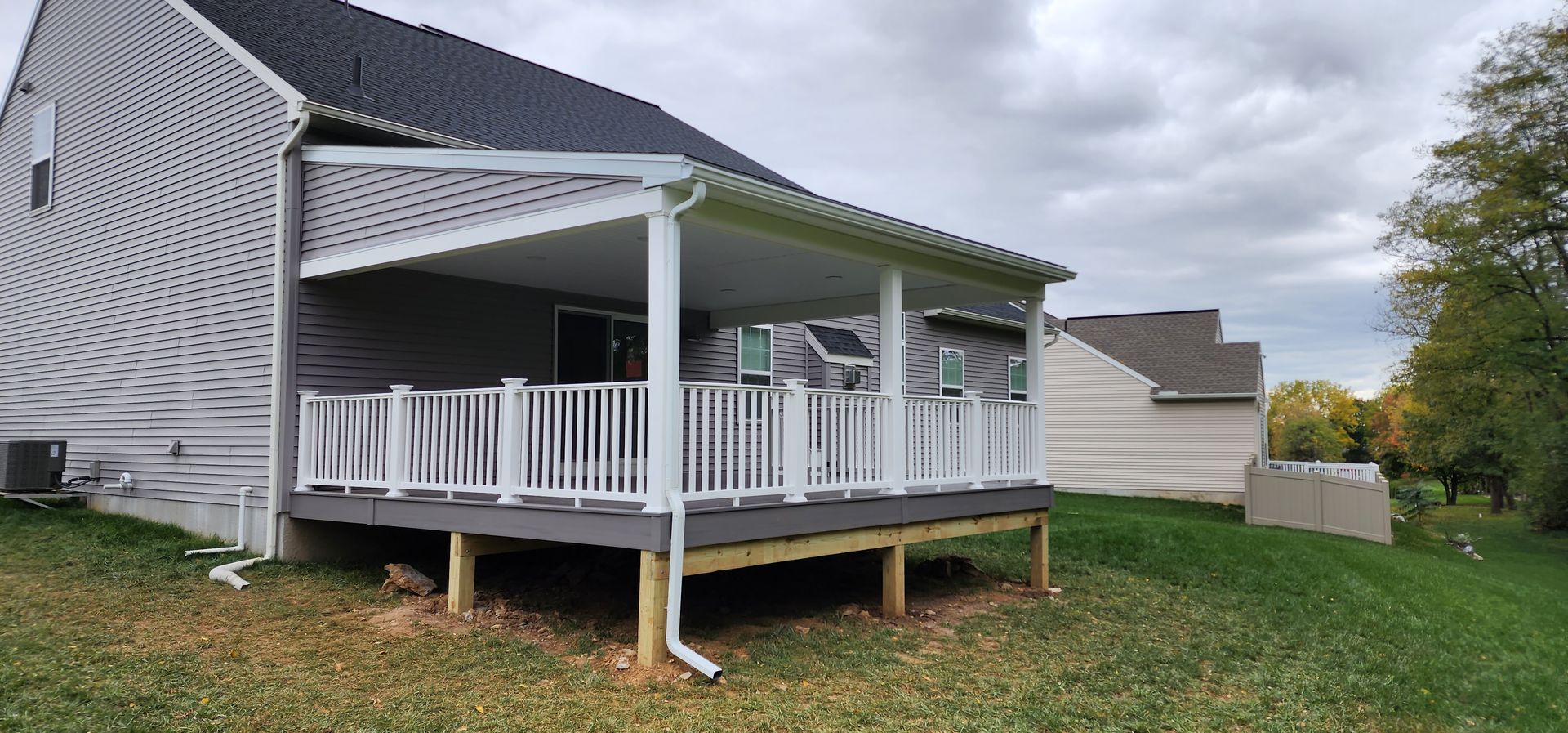 A gray-sided house features a elevated deck with white railings and a covered patio area set against a cloudy sky.