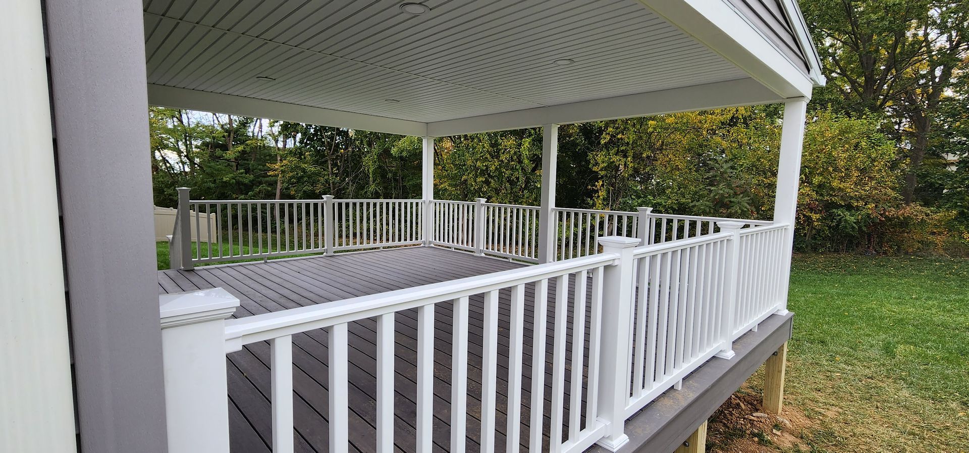 A covered wooden deck with white railings and support posts, overlooking a backyard with trees and grass.