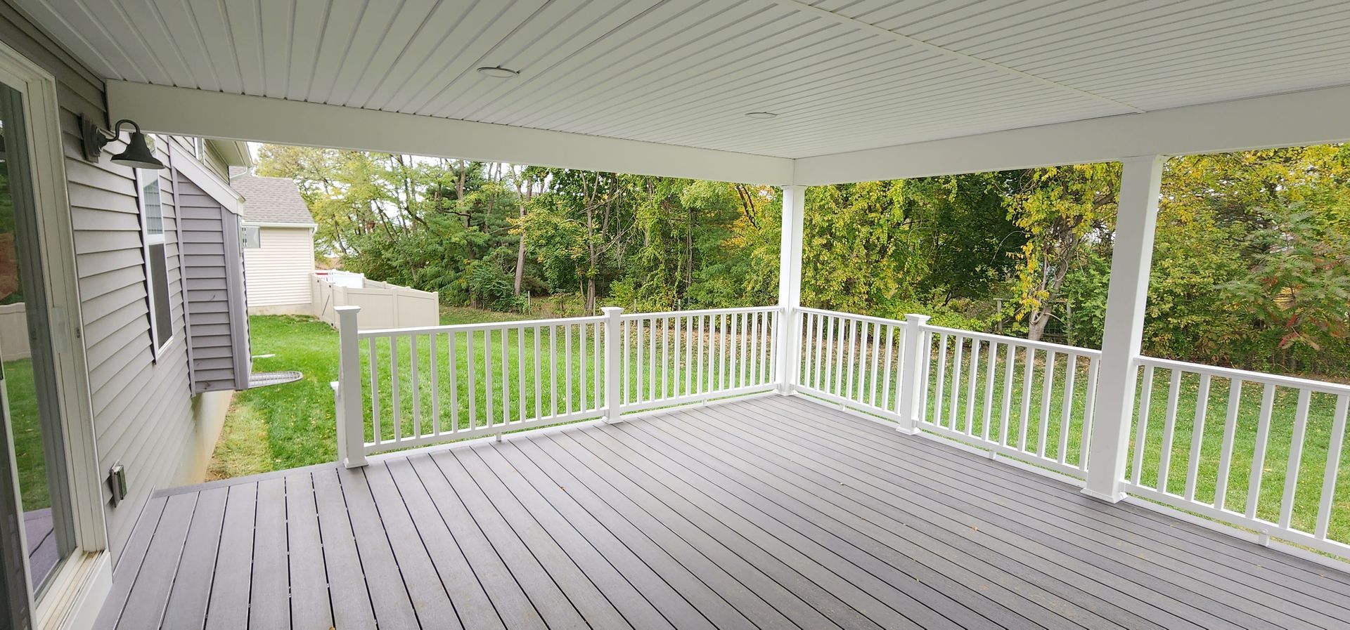 A gray wooden deck with a white railing and covered ceiling, overlooking a grassy yard with trees in the background.
