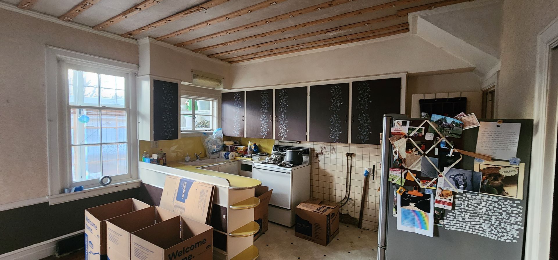 A cluttered, older kitchen undergoing renovations with exposed ceiling rafters, cardboard boxes, and dark cabinets.
